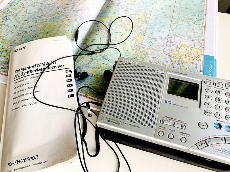 Photo of a Sony ICF-SW7600GR shortwave radio on a table. Next to the paperback book size radio are its instruction book, earbud headphones and a map.