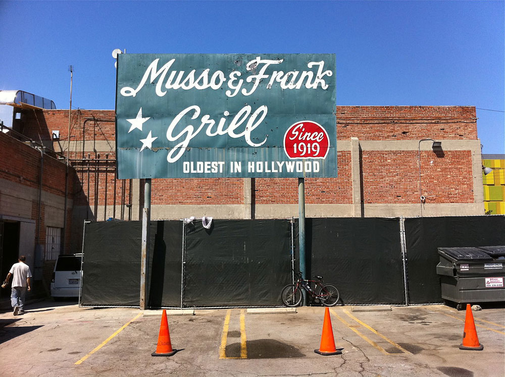 Photo taken of a large sign for Musso & Frank Grill in Hollywood on a bright sunny day. The dark green sign features the restaurant's name in white cursive lettering.