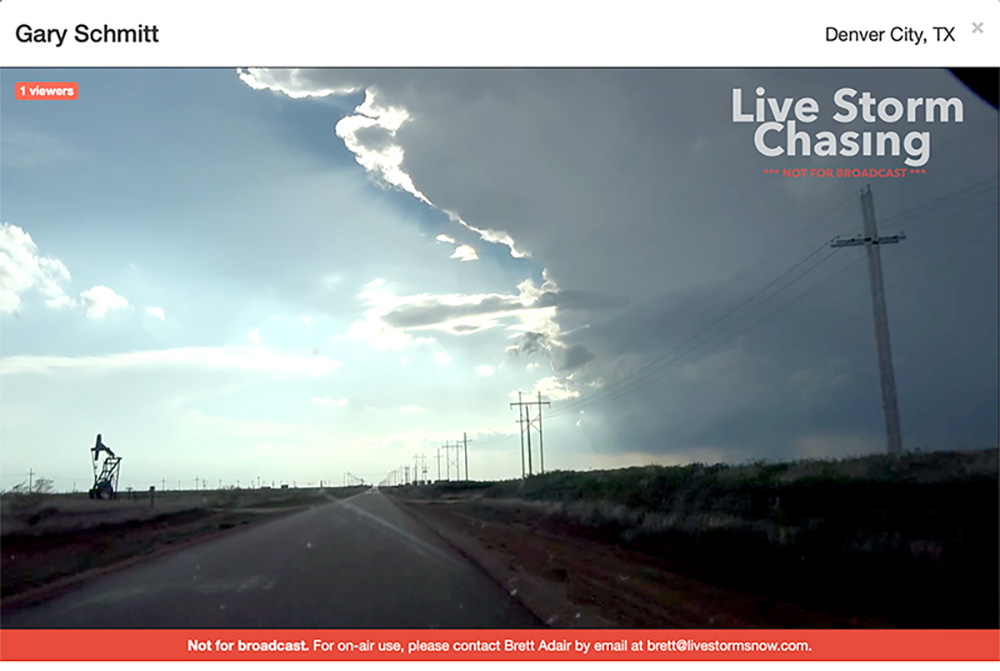 Screenshot from a live video feed at Live Storm Chasers. It shows a view of Texas flatlands as a gray thunderstorm brews on the horizon.