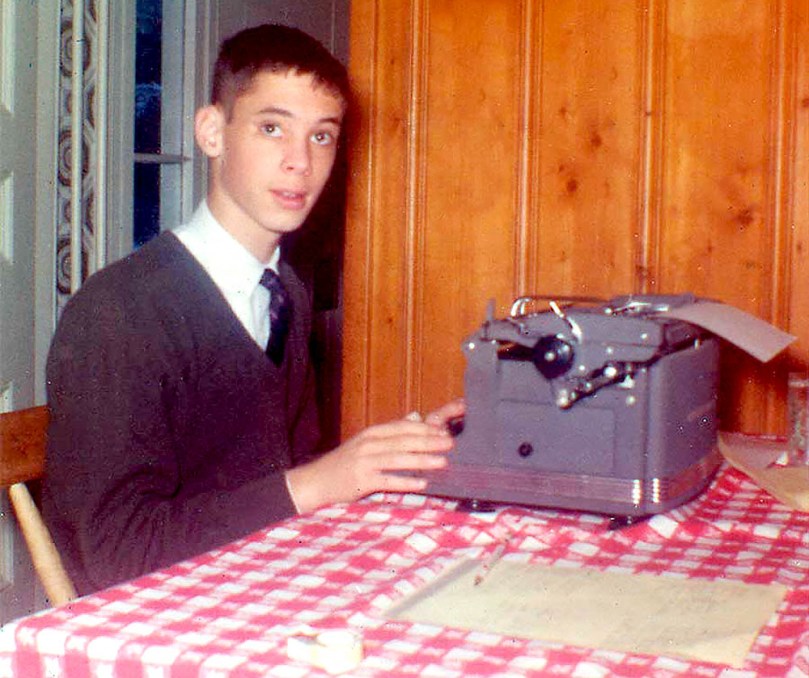 A young John Waters sits at a typewriter in what appears to be a table in a home's kitchen.