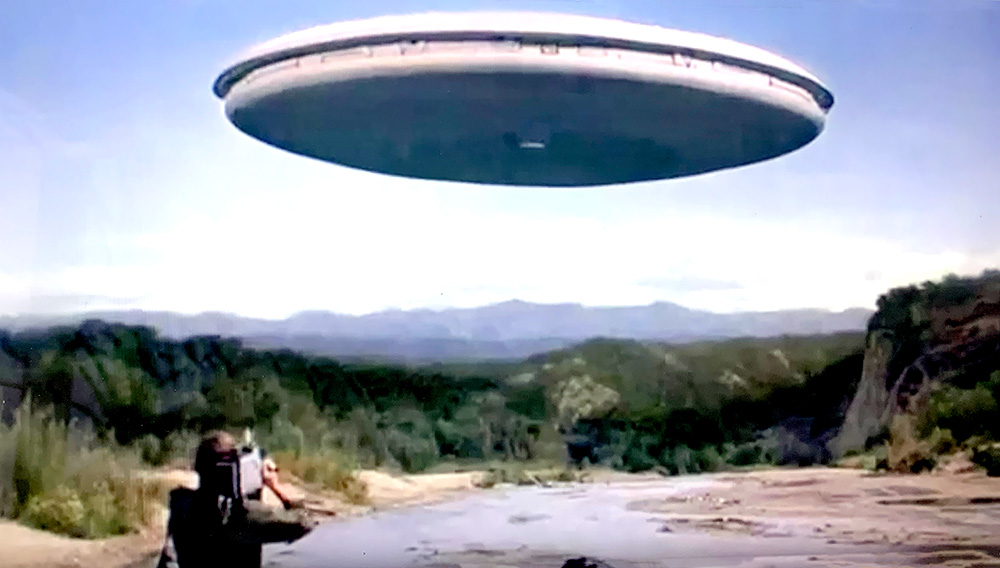 Journalist Marc Singer stands with his back to us as he videotapes a giant alien flyinf saucer gliding over a river in El Salvador.